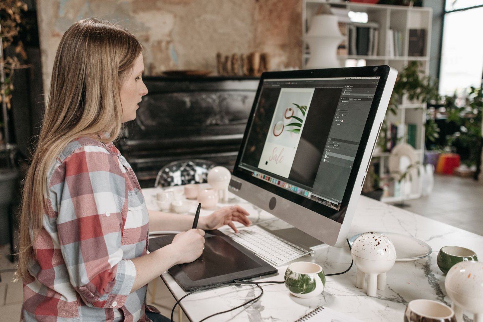 A designer at a large monitor with a print layout half assembled on screen, printed proofs weighted down on the desk beside